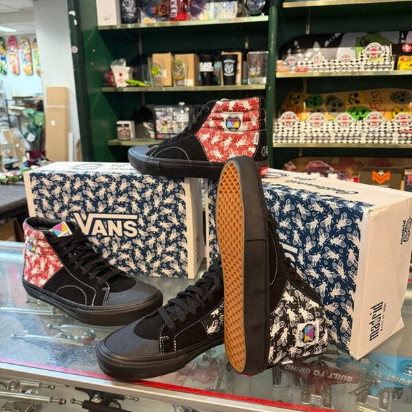 A pair of black Vans high-top sneakers with red-and-white patterned side panels, displayed on a glass counter in a retail store alongside matching Vans shoeboxes.