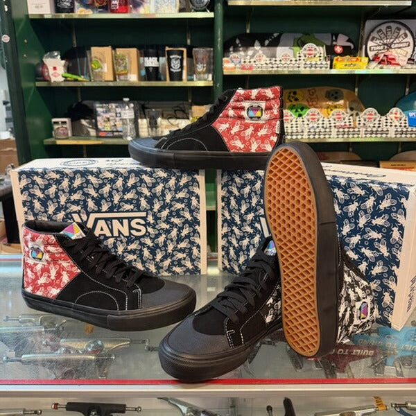 A pair of black Vans high-top sneakers with red and white patterned side panels, displayed on a glass counter in a retail store alongside their branded shoeboxes and skateboards.