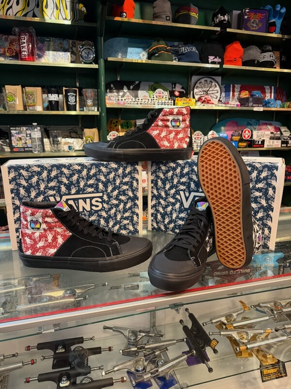 A pair of black high-top Vans sneakers with red and white patterned side panels, displayed on a glass counter in a retail store. The Vans shoeboxes are stacked behind them, and skateboards and other store merchandise are visible in the background.