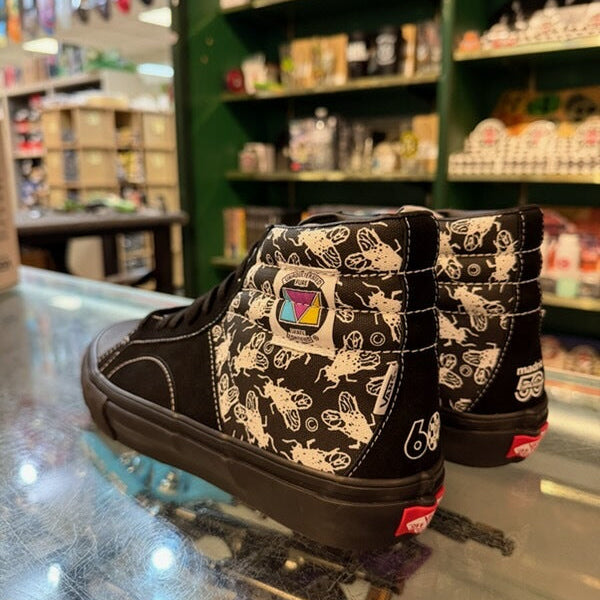 A pair of black high-top sneakers with a beige bee pattern and a colorful square patch on the side, displayed on a glass counter inside a retail store.