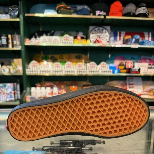 A single Vans shoe shown upside down, displaying its classic brown waffle-patterned rubber sole and black fabric upper, set against a blurred background of a skate shop interior.