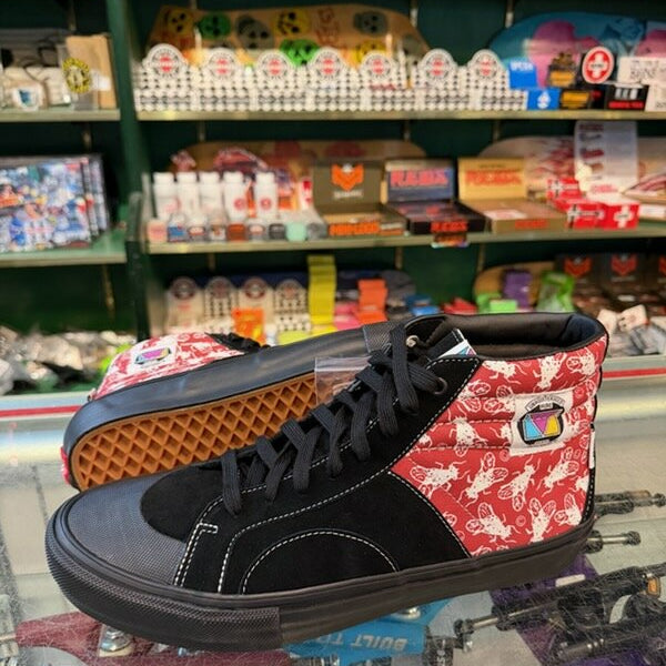 A pair of high-top sneakers displayed on a glass counter inside a retail store. The shoes feature a black suede and canvas upper with a red patterned side panel showing white horse silhouettes, black laces, and a waffle-patterned outsole.
