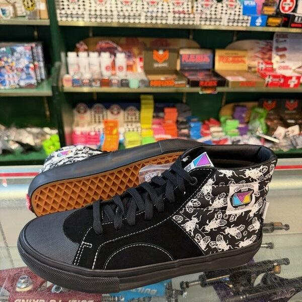 A pair of high-top skate shoes displayed on a glass counter inside a retail store. The shoes feature a black suede and canvas upper with a black-and-white graphic pattern on the side panels and a waffle-patterned orange outsole.