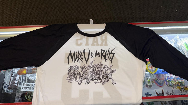 A raglan baseball t-shirt with a cream-colored body and black sleeves, featuring a black graphic print on the front with stylized text and an illustration. The shirt is laid flat on a hanger inside a retail display case.