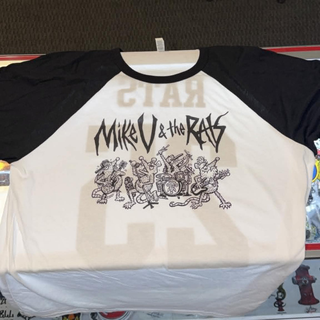 A black and white raglan baseball t-shirt featuring a black graphic print of the band 'Mikro & the Ras' with a cartoon character, displayed on a hanger on a store counter.