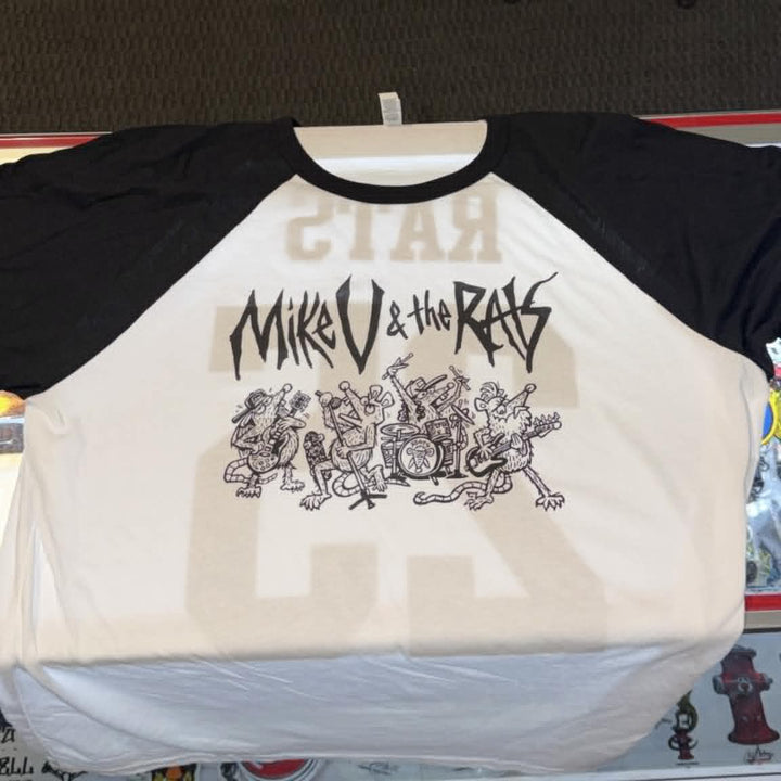 A black and white raglan baseball t-shirt featuring a black graphic print of the band 'Mikro & the Ras' with a cartoon character, displayed on a hanger on a store counter.