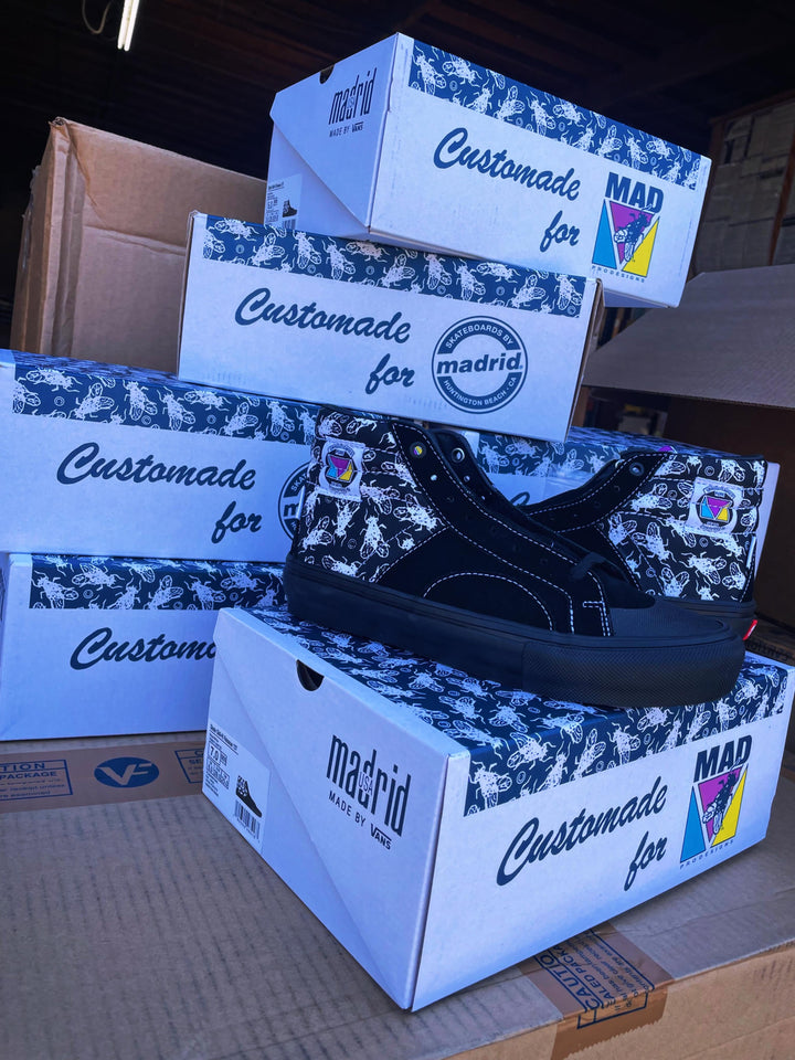 A black low-top sneaker with white contrast stitching and a colorful Vans-style heel patch, displayed on top of several white and blue 'Custommade for madrid' shoe boxes stacked on cardboard in a warehouse-like setting.
