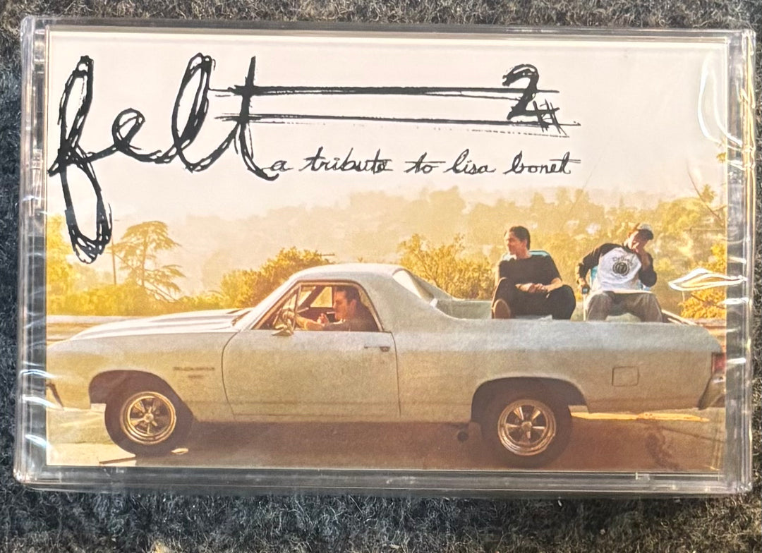 A clear plastic cassette case containing a music release titled 'felt 2' with the subtitle 'a tribute to lisa bonit'. The cover art shows a vintage white pickup truck on a warm-toned road with two people sitting in the bed, photographed on a dark textured fabric background.