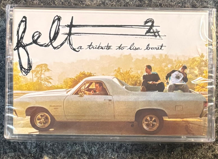 A clear plastic cassette case containing a music release titled 'felt 2' with the subtitle 'a tribute to lisa bonit'. The cover art shows a vintage white pickup truck on a warm-toned road with two people sitting in the bed, photographed on a dark textured fabric background.