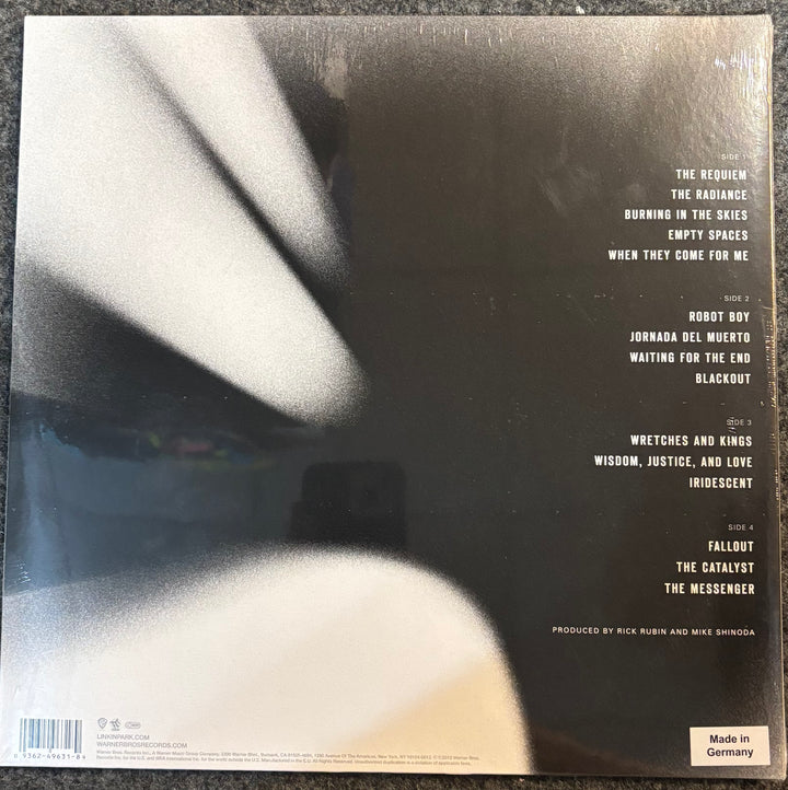 The back cover of Linkin Park - A Thousand Suns, featuring a black background with white text listing track titles and artist credits, photographed on a dark textured surface.