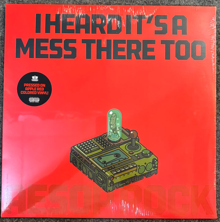 A vinyl record album cover for 'I Heard It's A Mess There Too' by Aesop Rock, featuring a bright red background with bold black text and an illustration of a green and brown control panel with a glowing green tube. The record is sealed in plastic shrink wrap and photographed flat on a dark textured surface.