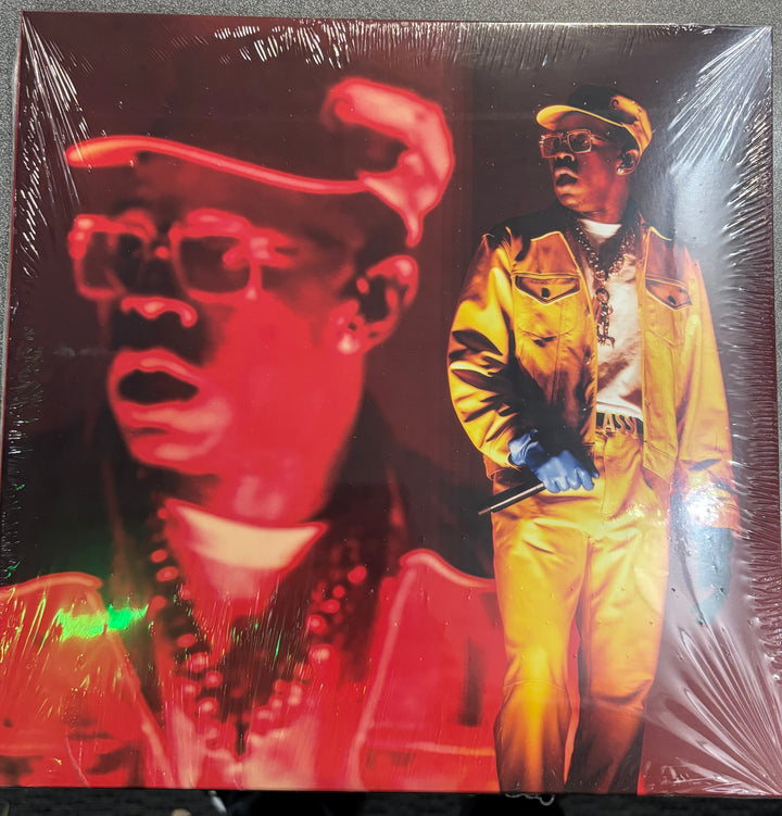 TYLER, THE CREATOR - LIVE AT LOLLAPALOOZA record with a cover featuring a person in a yellow jacket and another in red, on a reflective surface.