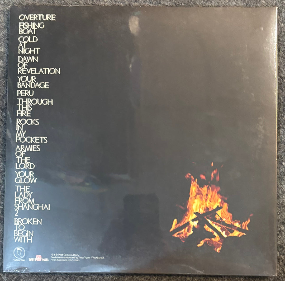 The back cover of a vinyl record sleeve for Mountain Goats - Through This Fire Across From Peter Balkan, featuring a dark black background, white text listing track titles, and a small burning fire graphic in the bottom right corner.