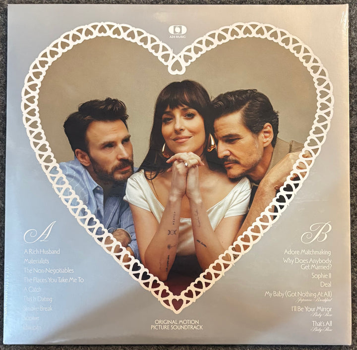 A square vinyl record cover for Materialists original motion picture soundtrack, featuring a heart-shaped white lace border framing a photo of the three main characters. The background of the cover is a warm beige with white typography listing the movie songs and tracks.