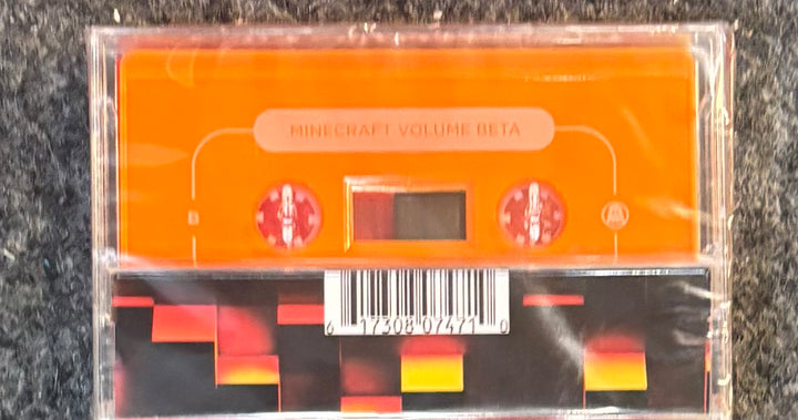 A cassette-style album cover for 'Minecraft Volume Beta' in a clear plastic sleeve, featuring an orange top half and a black bottom half with red and yellow geometric blocks, photographed on a dark textured surface.