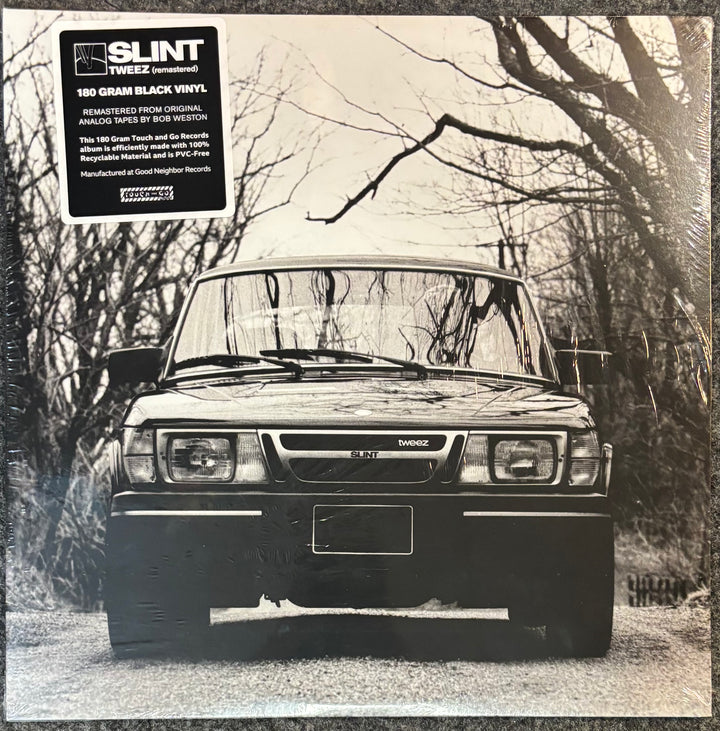 A vinyl record sleeve for Slint's 'Tweez' 180g black vinyl release. The cover features a black-and-white photograph of a car parked on a gravel road in a wooded area, with a black text box in the top-left corner containing track and artist details.