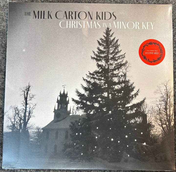 The Milk Carton Kids 'Christmas in a Minor Key' vinyl record cover, featuring a snowy winter scene with a church and a decorated Christmas tree. A red 'Festive Red' sticker is visible on the top right.