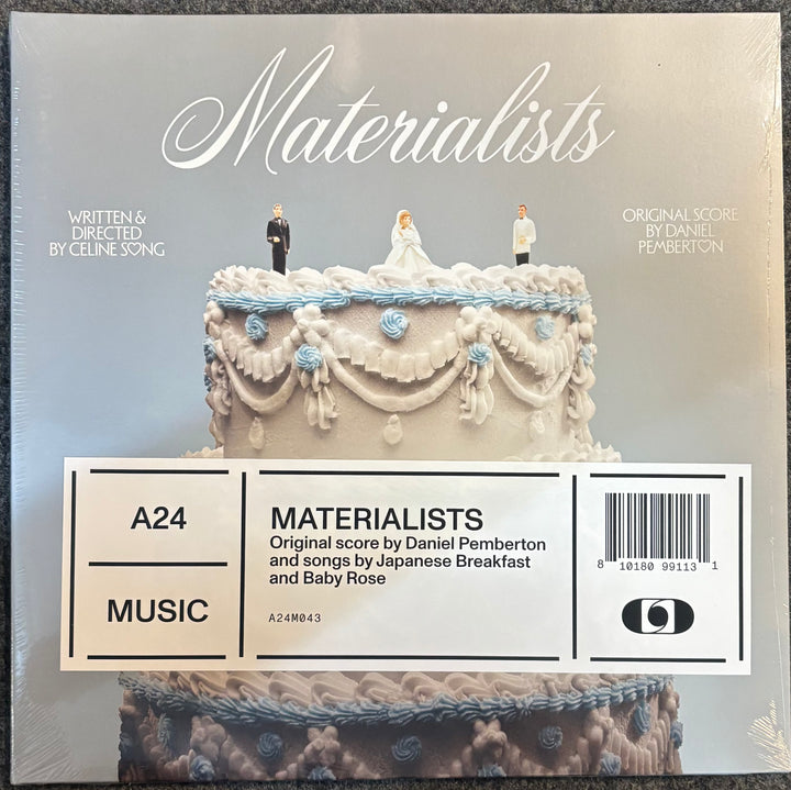 A24 Music 'Materialists' soundtrack album cover, featuring a white wedding cake with miniature bride and groom figures on top, with a white barcode label overlay at the bottom.