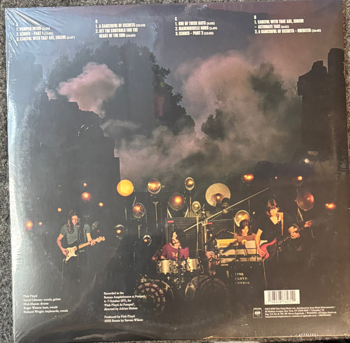 Pink Floyd – Pink Floyd At Pompeii-Mcmlxxi record cover with a band performing on stage under a dark sky with clouds.