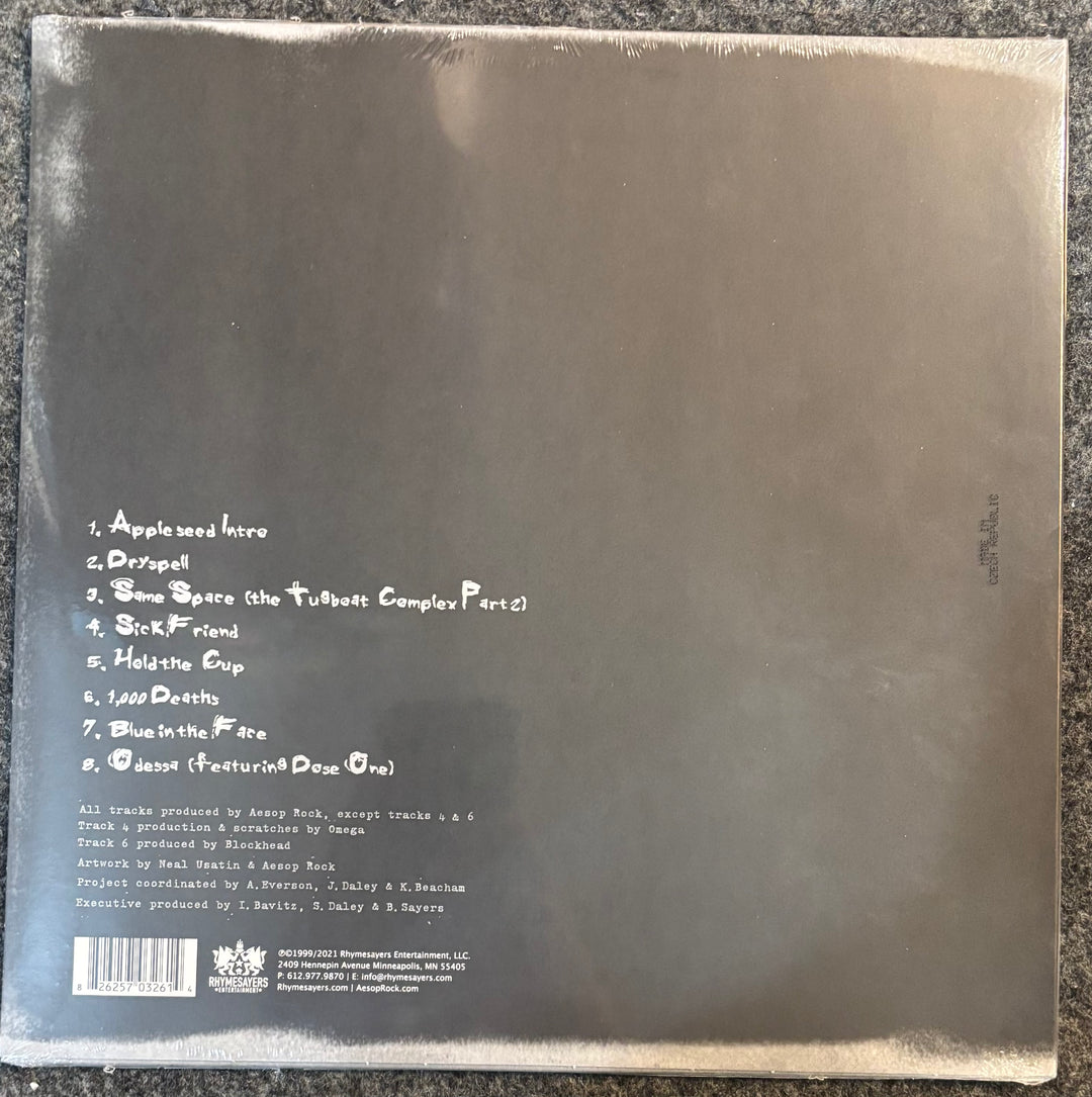 The back cover of Aesop Rock – Appleseed, photographed flat on a dark textured surface. The cover is dark gray with white handwritten-style text listing the tracks and credits, and a barcode in the bottom-left corner.