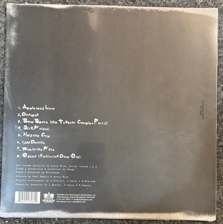 The back cover of Aesop Rock – Appleseed, photographed flat on a dark textured surface. The cover is dark gray with white handwritten-style text listing the tracks and credits, and a barcode in the bottom-left corner.