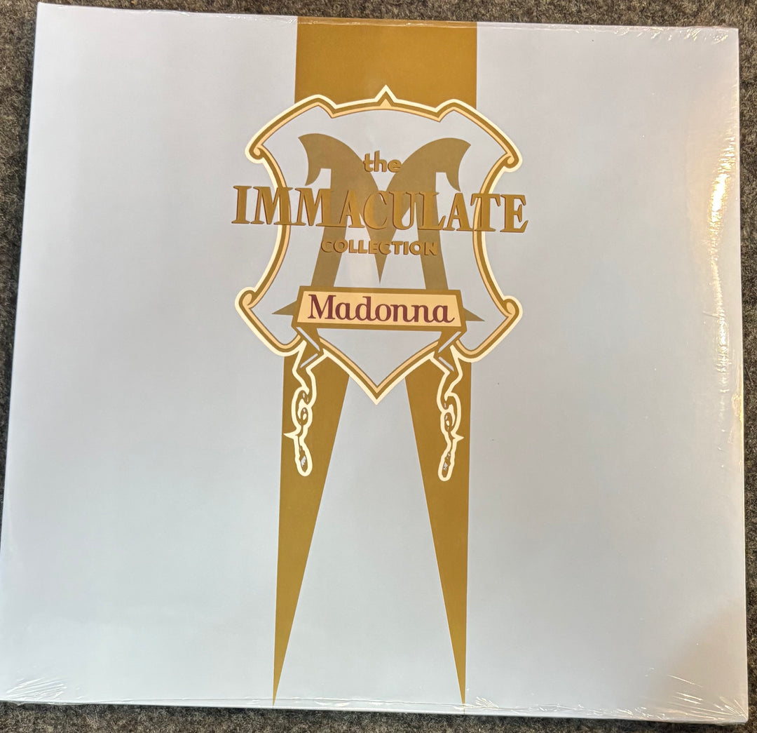 The front cover of Madonna's 'The Immolate Collection' vinyl record, sealed in plastic shrink wrap, photographed flat on a dark textured surface. The cover is white with a prominent vertical gold ribbon and a gold shield logo containing the text 'the IMMOLATE collection' and 'Madonna'.