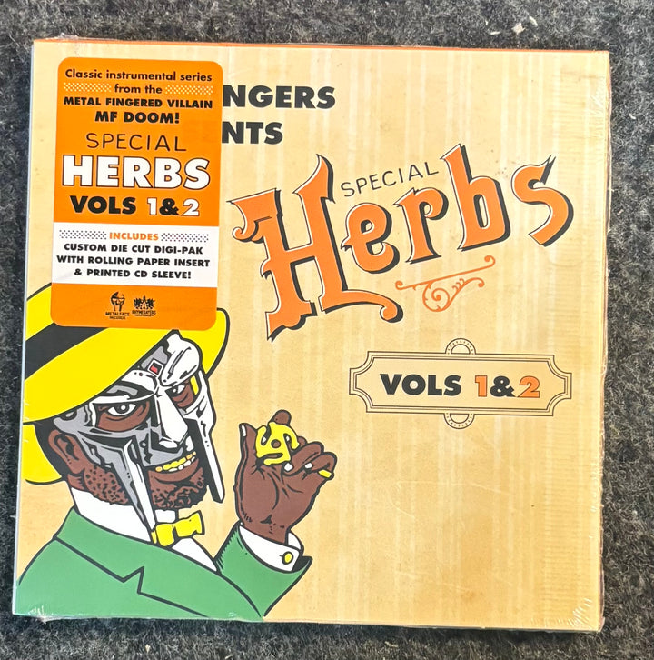Metal Fingers (MF DOOM) - Special Herbs Vol. 1 & 2. The packaging features a light wood-grain background with orange gothic lettering, an orange sticker on the left, and an illustration of a cyborg character in a yellow hat and green suit holding a small yellow mushroom.