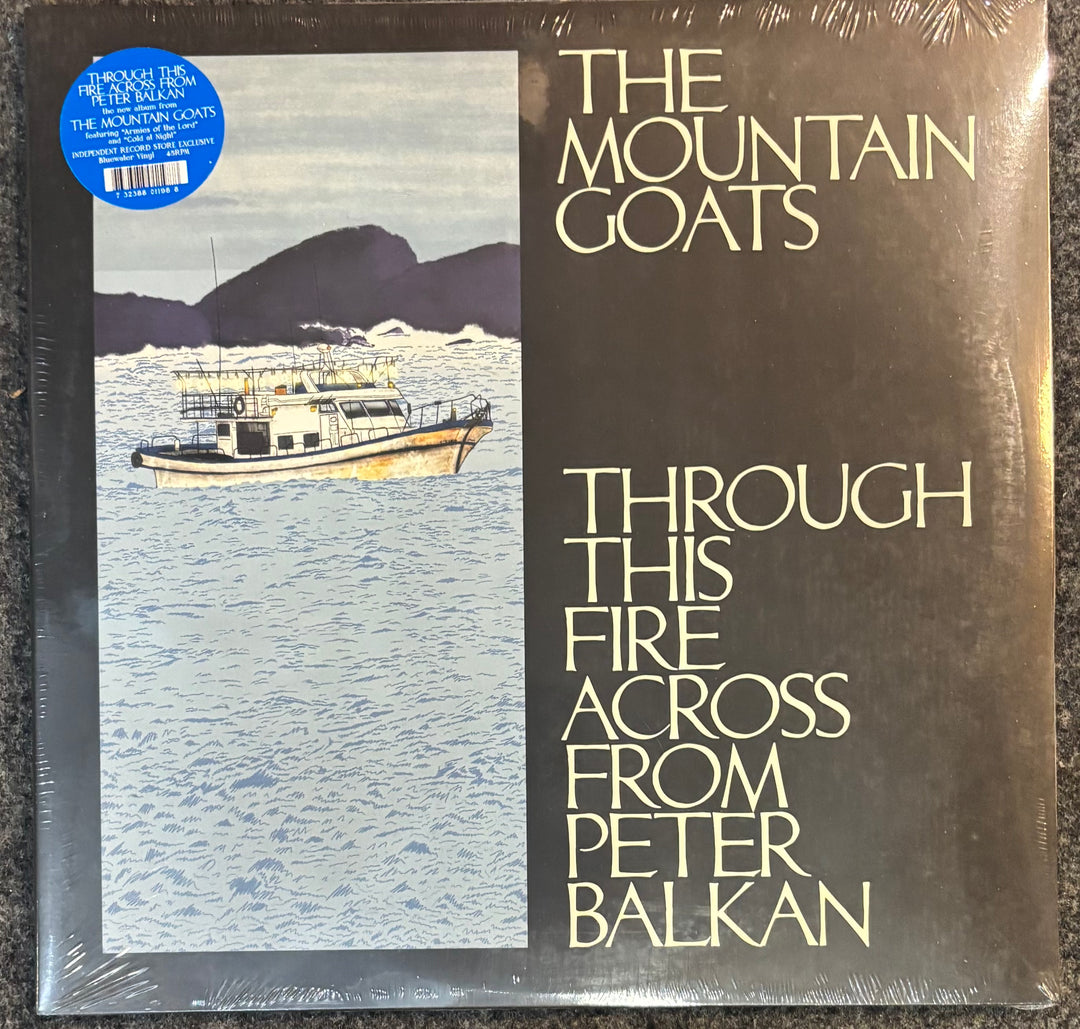Vinyl record album cover for Peter Balkan's 'The Mountain Goats / Through This Fire Across From', sealed in plastic with a blue sticker on the top left. The cover features a dark brown right side with cream text and a left panel showing a boat on blue water with dark mountains.
