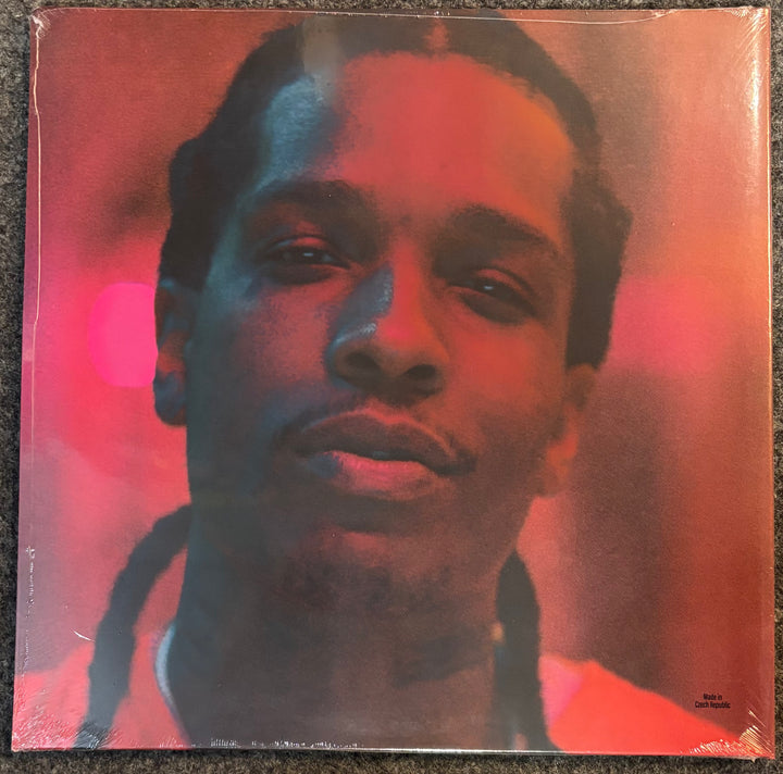 Highest 2 Lowest cover featuring a close-up portrait of a person with braided hair, overlaid with strong red and blue lighting effects. The cover is photographed flat against a dark textured surface, still wrapped in plastic.