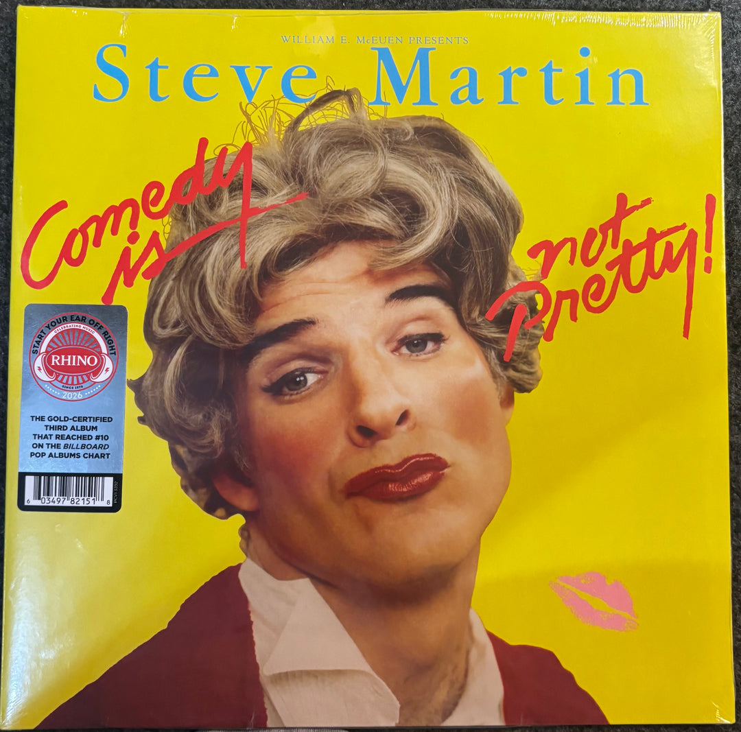 Steve Martin's 'Comedy Is Not Pretty!' vinyl record album cover, featuring a bright yellow background, a portrait of Steve Martin in a woman's hair and makeup, red handwritten text, and a Rhino 12-inch gold-plated LP sticker on the left.