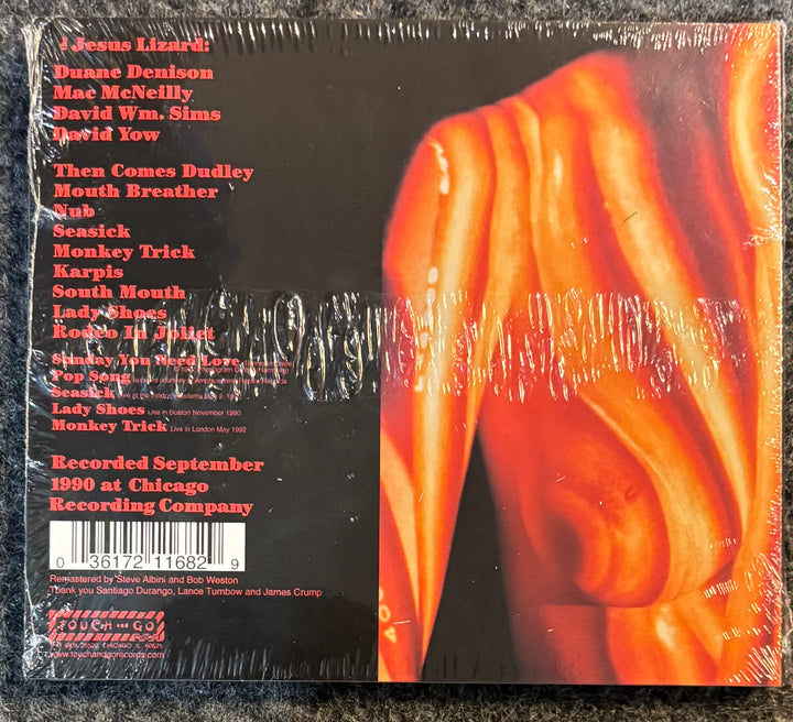 The back cover of The Jesus Lizard -Goat, still sealed in its plastic shrink wrap, showing a tracklist on a black background with a large orange abstract graphic on the right.