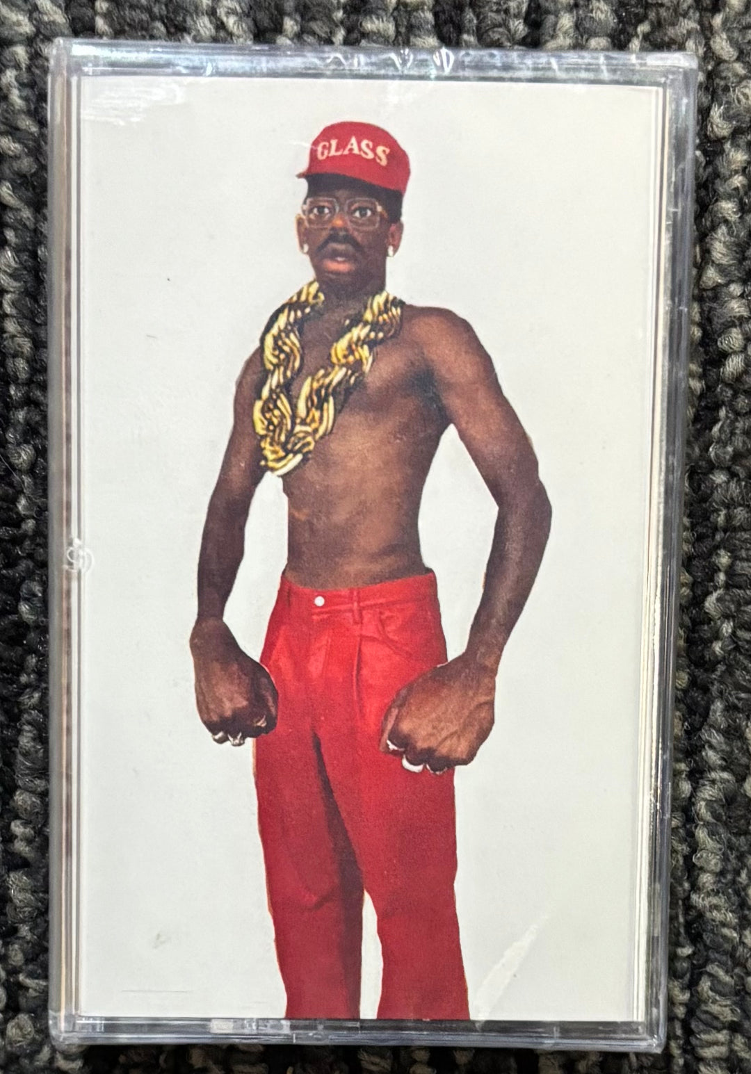 Tyler the Creator Don't Tap the Glass Cassette tape with a graphic of a person wearing a red cap and red pants on a textured surface.