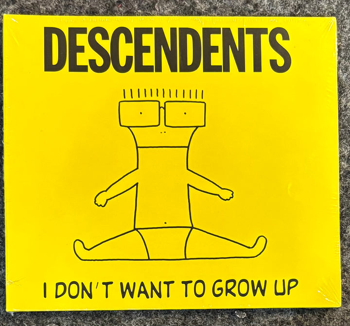 A square yellow album cover for the band Descendents, featuring a simple black line drawing of a seated figure and the text 'DESCENDENTS' at the top and 'I DON'T WANT TO GROW UP' at the bottom, photographed on a gray textured surface.