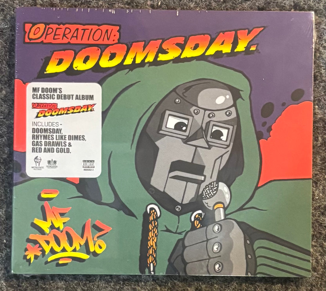 MF Doom - Operation: Doomsday, sealed in its plastic shrink wrap, placed on a dark textured carpet. The cover art features a stylized green hooded figure with a metal face mask holding a microphone, with bold red and yellow title text.