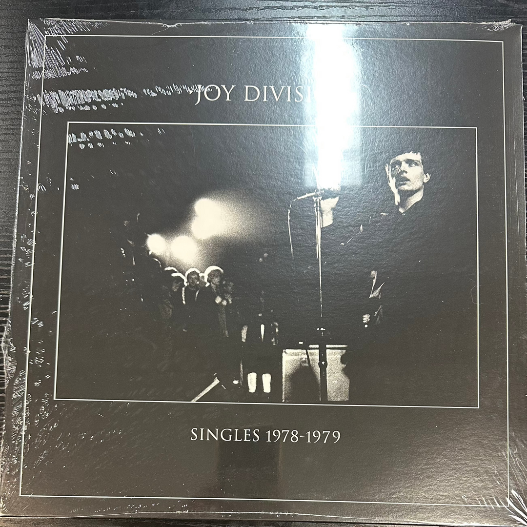 Joy Division album 'Singles 1978' on a vinyl LP record, with a black and white cover featuring a photo of the band performing.