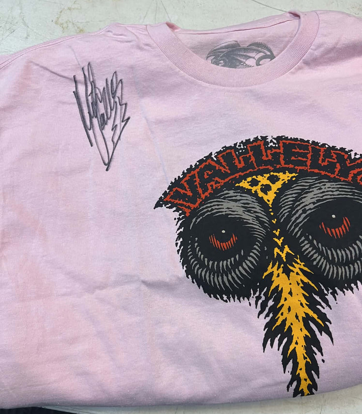 A light pink t-shirt featuring a large graphic of a stylized owl with black and yellow feathers and red eyes, under the word 'VALLEYS' in red. A black handwritten signature is printed on the upper left chest.