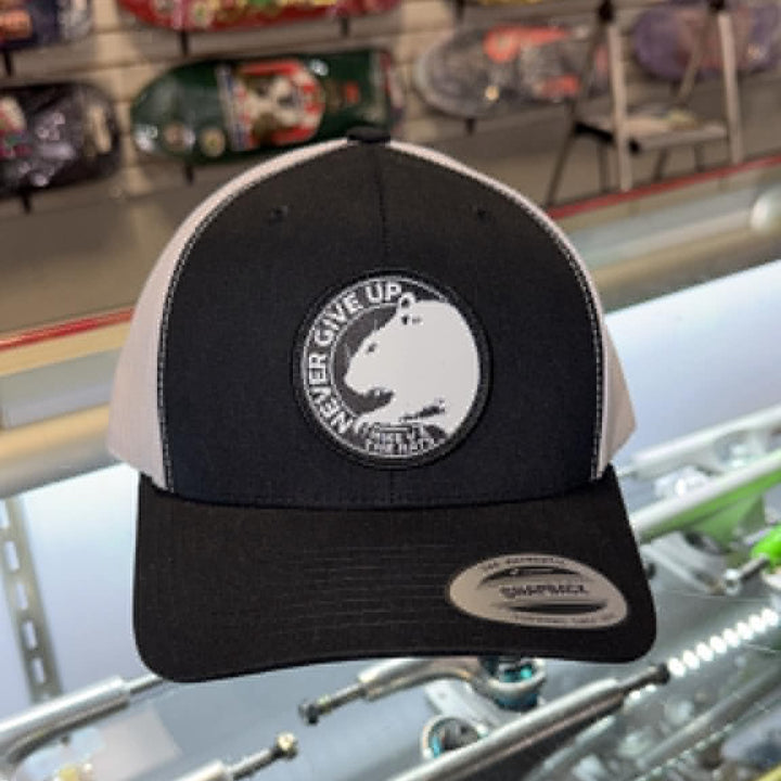 A black and white trucker hat with a circular patch reading 'GIVE US A CHANCE' and a bear graphic, displayed in a skate shop with skateboards in the background.