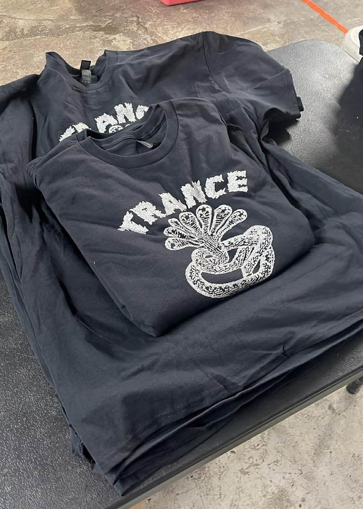 Black t-shirts with 'TRANCE' design on a folding table outdoors.