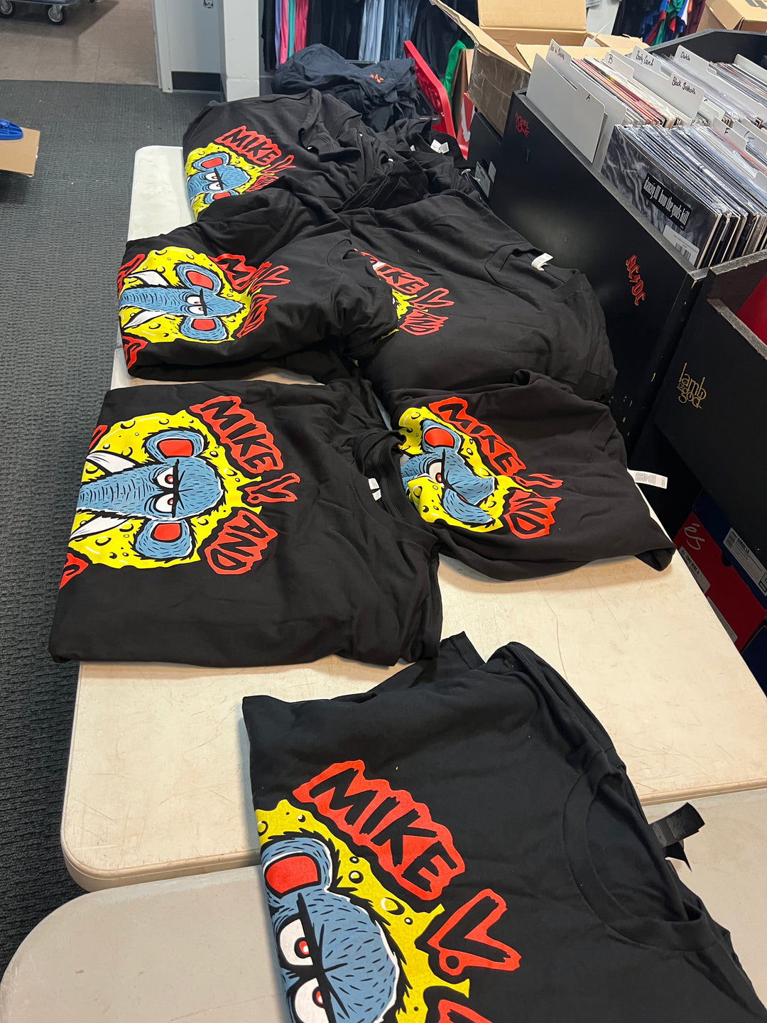 Several black graphic T-shirts laid out on a table in a storage or retail space. The shirts feature a large cartoon-style blue character on a yellow background with red and orange text reading 'MIKE V.